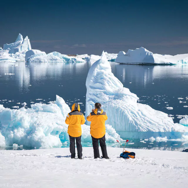 Zwei Passagiere stehe an der Küste der Antarktis und schauen auf das Meer mit Eisbergen.